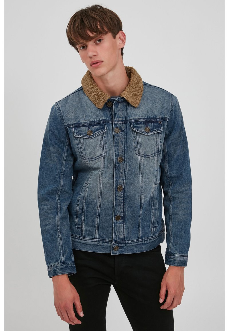 Denim jacket in faded blue with a brown sherpa collar. Features two chest pockets and button closure. Textured fabric with visible stitching.