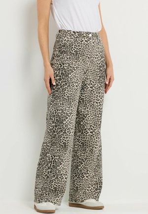 Frau trägt hoch taillierte, weit geschnittene Hosen mit beigem und schwarzem Leopardenmuster, kombiniert mit weißen Turnschuhen und einem weißen Oberteil.