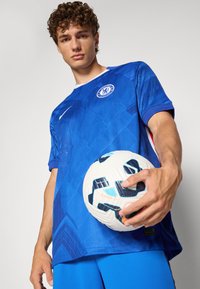 Blå fodboldtrøje med geometrisk tekstur og logo, parret med en hvid og mønstret fodbold, der holdes i venstre hånd.