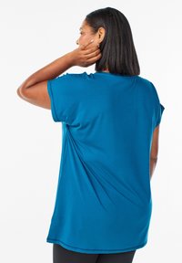 Tirkīza īsām piedurknēm blūze ar brīvu piegriezumu, gludu tekstūru un sānu šķirbēm, valkāta ar melnajām legingiem. Skats rāda aizmuguri un plecu detaļas.