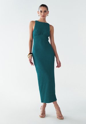 Robe côtelée teal avec une silhouette droite, un design sans manches et une taille froncée. Associée à des talons beiges à lanières et des accessoires.