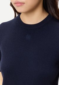 Ung person som bär en marinblå kortärmad stickad tröja med en liten broderad emblem centrerad under halsringningen.