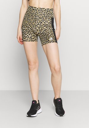 Žena nosící sportovní šortky s vysokým pasem s leopardím potiskem, černými bočními pruhy a černé tenisky Adidas s růžovými logy.