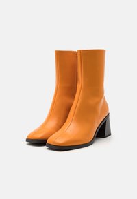Botas de tobillo de cuero naranja con punta cuadrada, textura suave y tacón grueso negro. Presentan una cremallera lateral para ponerlas y quitarlas fácilmente.