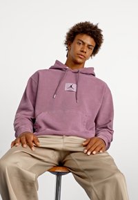 Sudadera morada con un bolsillo frontal, cordones y un parche con un logo. Llevada con pantalones beige, sentado en un taburete contra un fondo liso.