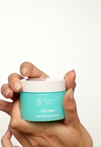 En turkos burk märkt "STAY WELL TEA TREE Light Calming Cream" som hålls i en hand, med ett vitt lock och en slät textur.