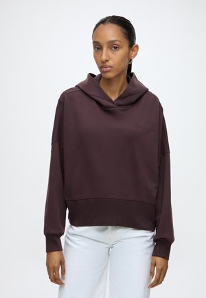 Femme aux cheveux raides et foncés porte un sweat à capuche ample marron foncé et un pantalon blanc, debout devant un fond clair uni.
