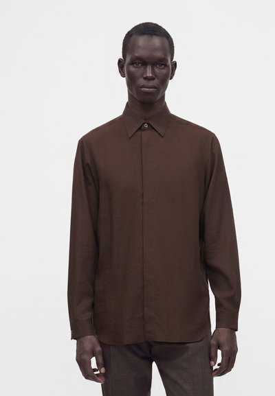 Homme portant une chemise marron foncé à manches longues boutonnée et un pantalon foncé, debout devant un fond clair uni.
