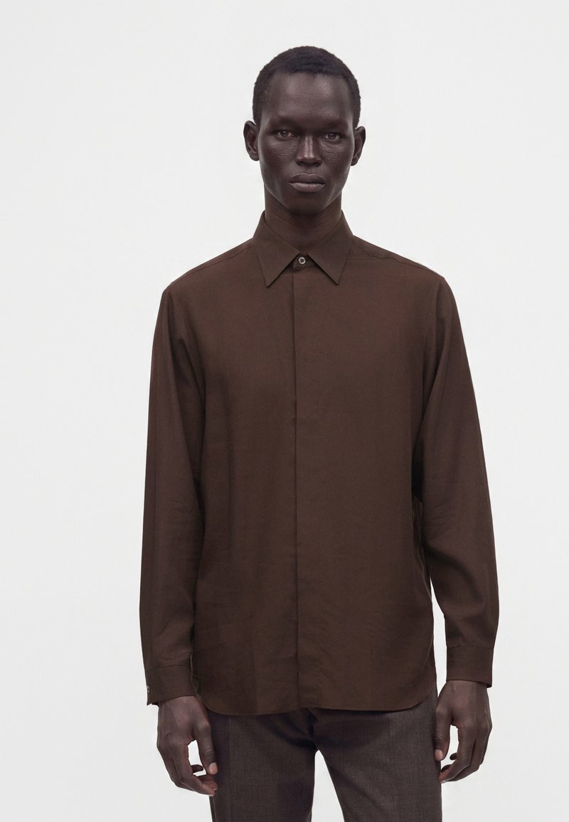 Homme portant une chemise marron foncé à manches longues boutonnée et un pantalon foncé, debout devant un fond clair uni.