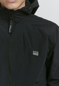Black windbreaker jacket with a zip closure, featuring a small, rectangular brand patch on the chest and a hood with adjustable drawstrings.