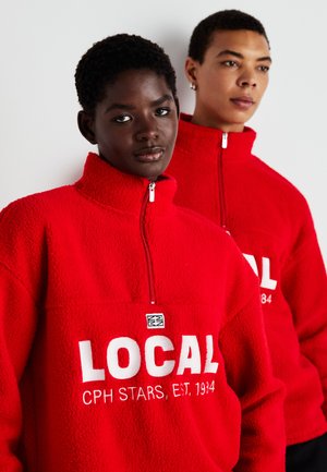 Red fleece zip-up jackets with a high collar, featuring bold white text "LOCAL" and additional branding details on the chest.