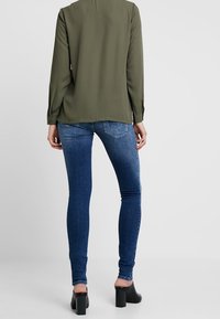 Olivengrøn langærmet bluse parret med blå skinny jeans og sorte blokhælede sko. Glat stof, casual snit og subtil glans.