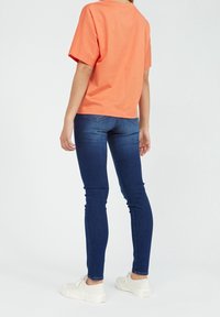 Krátké oranžové tričko s volným střihem a kulatým výstřihem, nošené s tmavě modrými skinny džíny a bílými teniskami.
