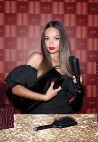 Lisseur à cheveux noir tenu à côté d'une pochette noire brillante. Le sujet a des cheveux raides et lisses, des lèvres rouges, et est positionné devant un fond rouge à motifs. Une brosse à cheveux noire avec une large tête se trouve sur une table dorée étincelante.