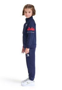 Completo felpa due pezzi navy con giacca con zip e pantaloni coordinati. Presenta un logo rosso sulla manica, dettagli bianchi, e abbinato a sneaker bianche.
