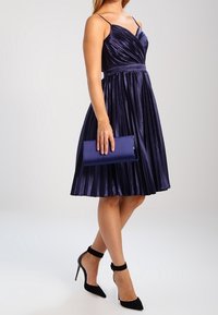 Fialové plisované šaty s těsně padnoucím živůtkem a tenkými ramínky, doplněné o odpovídající kabelku a černé sandály na kotníkovém pásku.