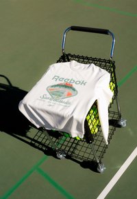 Felpa bianca Reebok con design da campo da tennis appoggiata su un carrello per palline da tennis su un campo esterno con linee verdi e bianche.