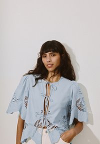 Lichtblauwe blouse met fluttermouwen, uitgesneden kanten details en striksluiting bij de halslijn. Zachte stof met een textuurafwerking.