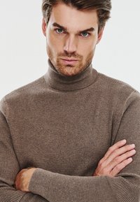 Brązowy sweter z golfem w prążki, wykonany z miękkiego materiału, o dopasowanym kroju i długich rękawach, noszony ze skrzyżowanymi ramionami.