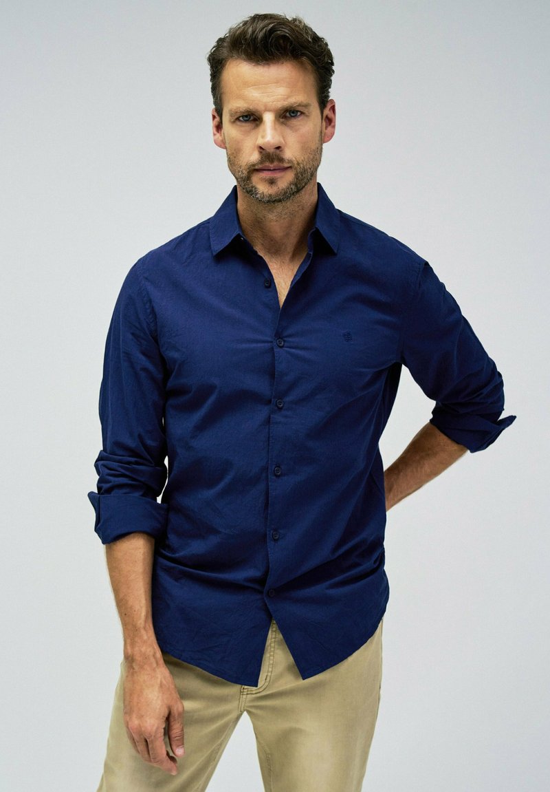 Camisa de hombre azul marino de botón en tejido texturizado; presenta un cuello clásico, mangas largas y un pequeño logo en el pecho.