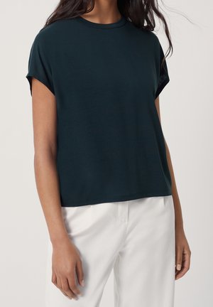 Frau trägt ein dunkelgrünes Kurzarmshirt mit umgeschlagenen Ärmeln und weiße Hose, steht vor einem hellen, schlichten Hintergrund.