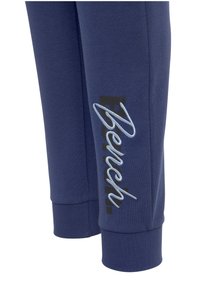 Marineblauwe joggingbroek met geribbelde boorden, voorzien van een zwart bedrukt logo en decoratieve lichtblauwe stiksel aan de zijkant.