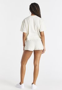 Kvinna med flätat hår iförd vit randig kortärmad topp och matchande shorts med vita sneakers, stående vänd bort från kameran mot vit bakgrund.