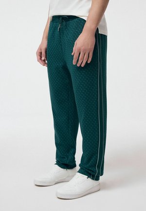 Mężczyzna ubrany w ciemnozielone spodnie dresowe w wzory z białymi paskami po bokach i białe trampki, stojący na jednolitym tle.