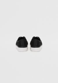 Baskets basses en mesh noir avec des semelles blanches, vues de dos sur un fond gris clair uni.