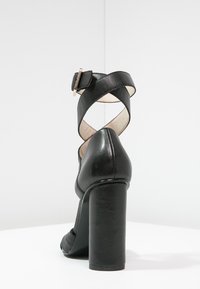 Musta nahkainen korkea sandaali, jossa on paksu korko ja ristikkäiset nilkkahihnat, jotka on kiinnitetty pienellä soljella, nähtynä takaa.