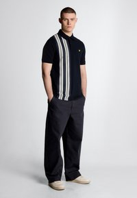 Polo navy con strisce verticali bianche, maniche corte e un emblema giallo. Abbinato a pantaloni scuri e sneaker chiare.