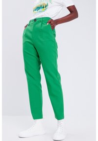 Pantalon vert en tissu lisse, avec une taille haute, des poches latérales et une fermeture par un seul bouton, associé à des chaussures blanches.