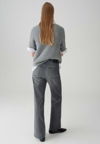 Vrouw met lang haar die een grijze fuzzy trui over een wit shirt draagt, grijze wijde jeans en zwarte platte schoenen, staand met de rug naar voren gericht.