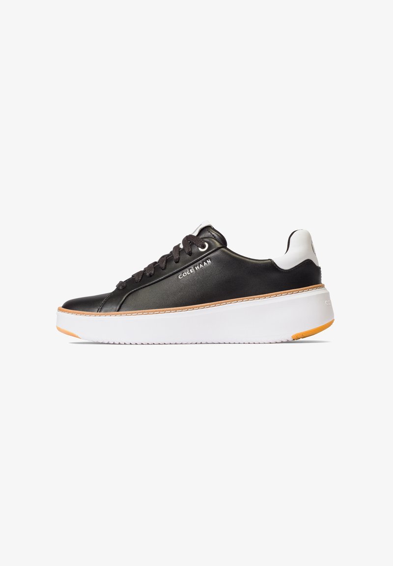 Zapatilla sintética negra con media suela blanca, detalle naranja en la suela exterior y cordones planos. Presenta un acabado liso y un branding sutil en el lateral.