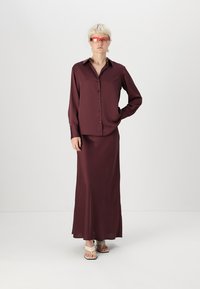 Bordová saténová blúzka s guľôčkovým zapínaním a dlhými rukávmi, skombinovaná s dlhou sukňou v rovnakom odtieni. Model nosí priehľadné, tónované slnečné okuliare a sandále.