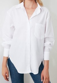 Camicia bianca a maniche lunghe realizzata in tessuto leggero, con colletto, chiusura a bottoni e un taschino sul petto. Design con orlo curvo.