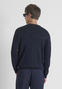 Marineblauwe trui met een ronde hals, geribde manchetten en gestructureerde schouderdetails, gecombineerd met donkere broek en rechthoekige zwarte zonnebril.