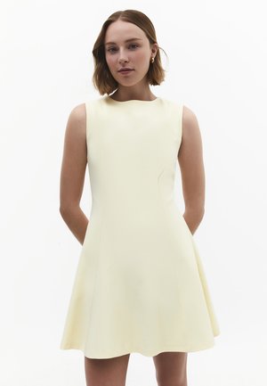 Femme aux cheveux bruns courts portant une robe sans manches jaune pâle, debout devant un fond blanc uni, les mains derrière le dos.