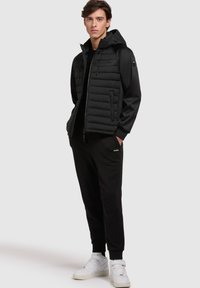 Zwarte gewatteerde jas met capuchon, met een gladde textuur en een rits aan de voorkant. Gecombineerd met zwarte sweatpants en witte sneakers.