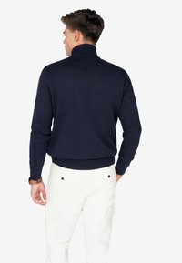 Maglione a collo alto blu navy realizzato in tessuto a maglia, caratterizzato da un design aderente con polsini e orlo a coste, indossato con pantaloni cargo bianchi.