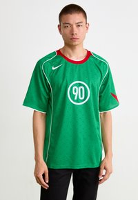 Zaļa sporta krekls ar sarkanām akcentēm, kurā centrā ir apaļš logo ar skaitli "90". Izgatavots no viegla, teksturēta auduma ar baltām apmalēm.
