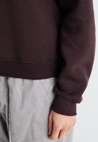 Maroon sweatshirt med ribbad nederkant och manschetter. Tyget ser mjukt ut, med en avslappnad passform. Kombinerad med ljusgrå byxor.