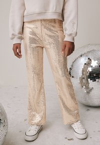 Pantaloni svasati in oro metallizzato decorati con strass, caratterizzati da una texture liscia e una finitura ad alto lucido, abbinati a sneakers bianche.