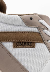 Gros plan sur le côté d’une basket montrant un tissu en mesh blanc, une bande en suède marron avec une étiquette "OMBRE" et du cuir synthétique beige avec des lacets blancs.