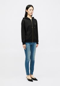 Casaco com fecho éclair preto, com um bolso frontal, capuz com cordão e detalhes em dourado, combinado com jeans skinny azuis e sapatos pretos de slip-on.