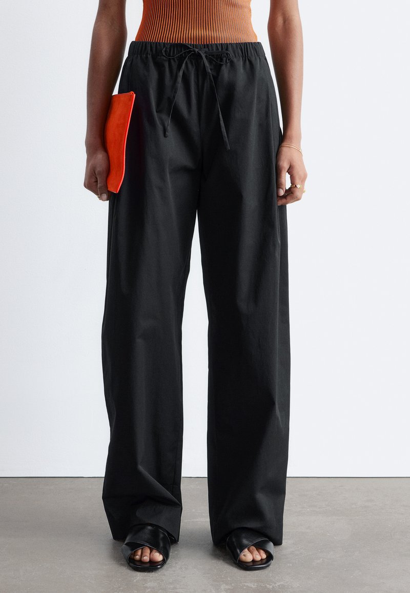 Woman wearing loose black drawstring pants, black open-toe heels, rust-colored ribbed top, holding small red clutch in left hand.