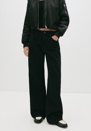 Person iført sort cropped bomberjakke, sorte spraglede vidde bukser og sorte og hvide loafers, stående med den ene hånd i lommen.