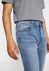 Hombre vistiendo jeans de mezclilla azul claro y una camiseta gris de manga corta, con los brazos relajados a los lados frente a un fondo blanco liso.