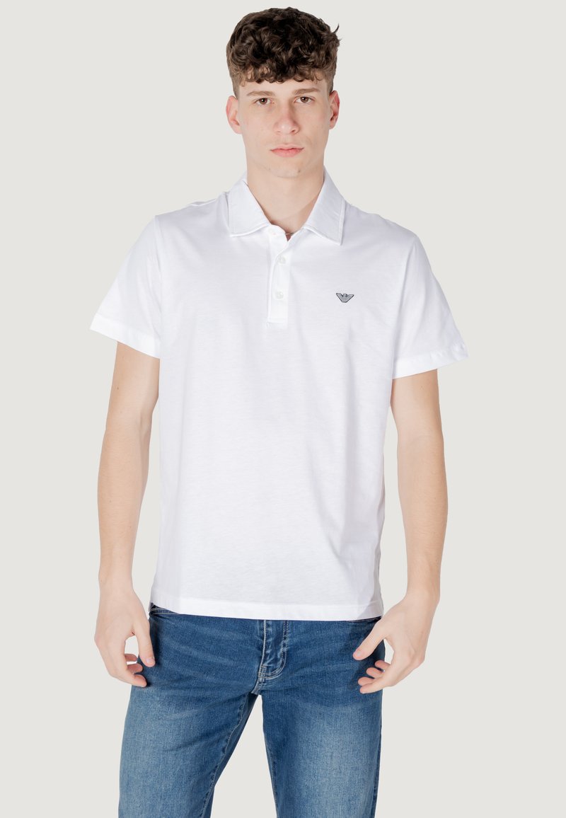 Polo shirt blanc en coton, avec un col classique, des manches courtes, et un logo brodé discret sur la poitrine. Porté avec un jean bleu.