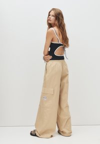 Top in cotone nero con schiena scoperta e dettagli bianchi; pantaloni beige a gamba larga con tasca cargo; modella con sandali e capelli lunghi sciolti.